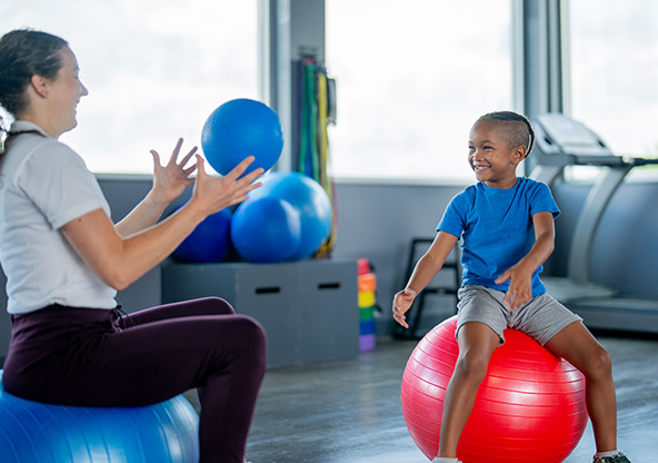 A young boy sits on an exercise ball and watches his occupational therapist throw a ball
